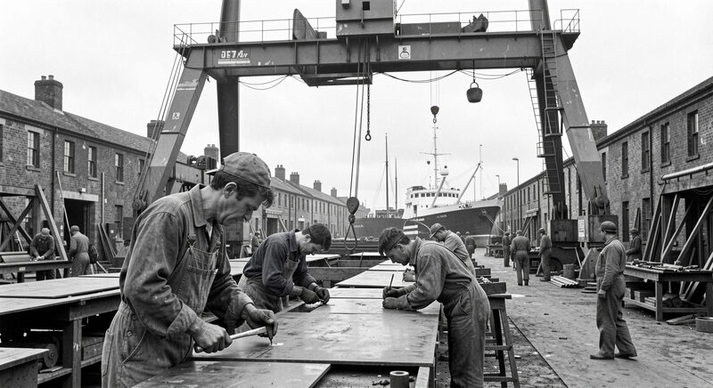 Shipyard workers in overalls riveting steel plates under a tall gantry crane in an old Clyde yard.